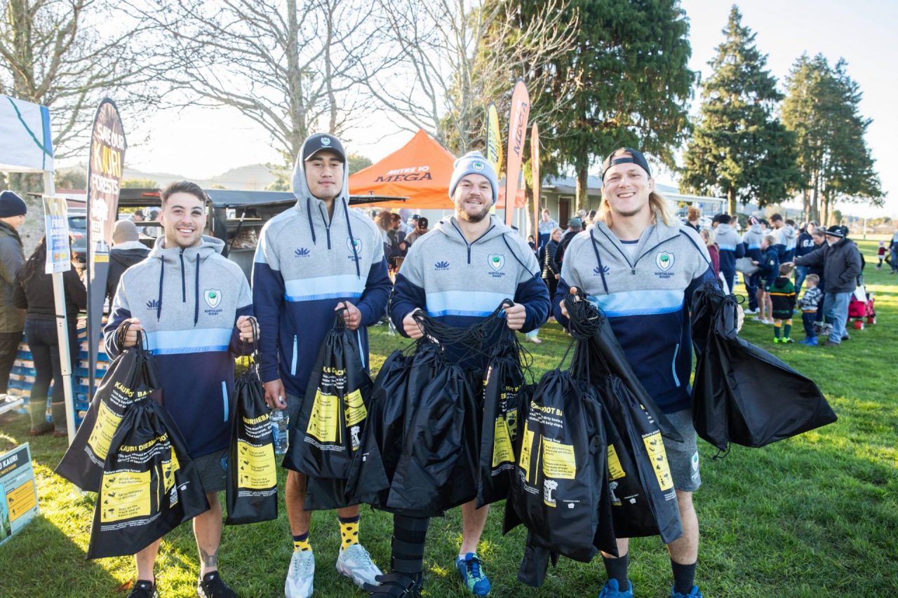 4 Northland Rugby players standing on Kensington Park holding Keep Kauri Standing boot bags