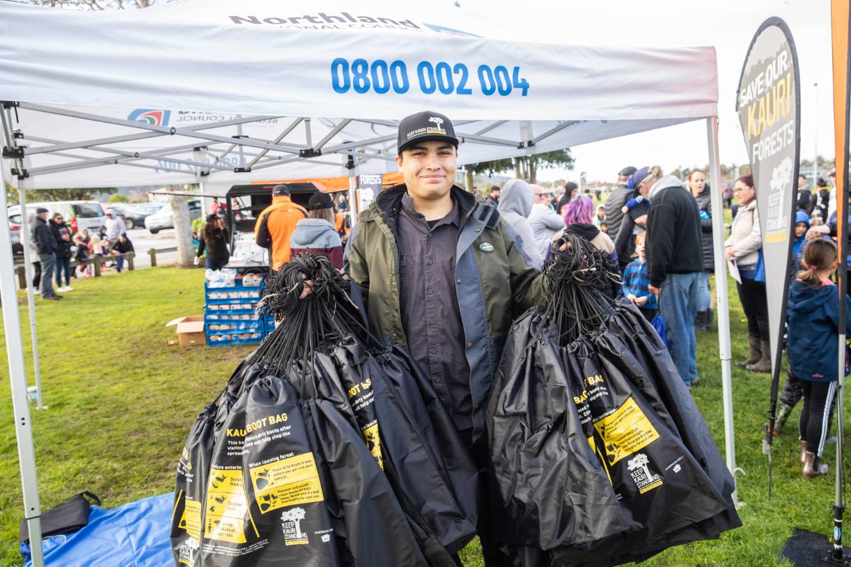 Man holding Keep Kauri Standing boot bags in front of Northland Regional Council tent at Kensington Park